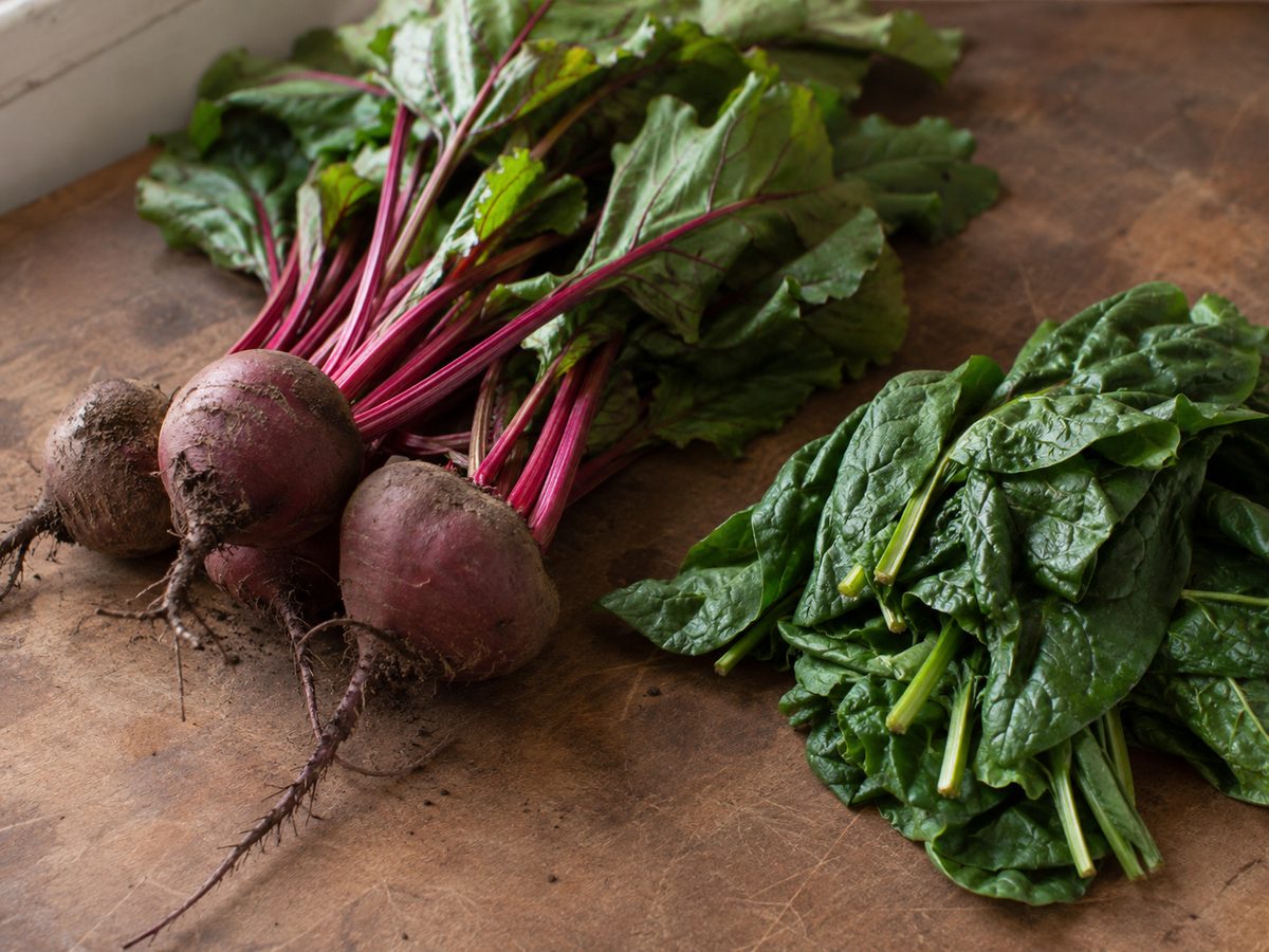 Fresh beetroot with leafy tops still attached, soil clinging to the skins, beside a pile of dark leafy greens on a worn wooden board
