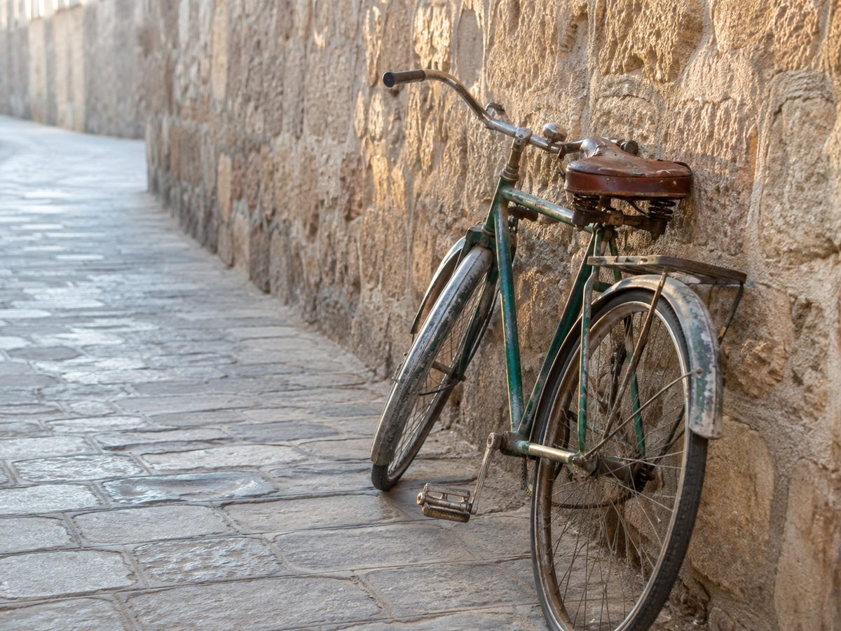 A well-worn green bicycle with a leather saddle leaning against a rough stone wall beside a cobbled lane in morning light