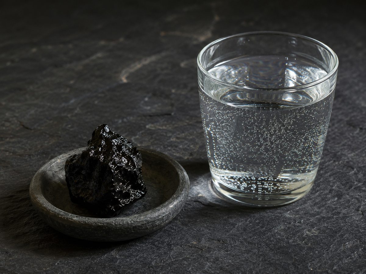 A piece of dark glossy shilajit resin in a small stone dish beside a glass of water with fine bubbles, on a dark slate surface