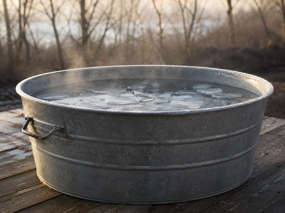 A galvanised metal tub of water with floating ice standing on weathered decking outdoors, mist rising from the surface and frost on the rim