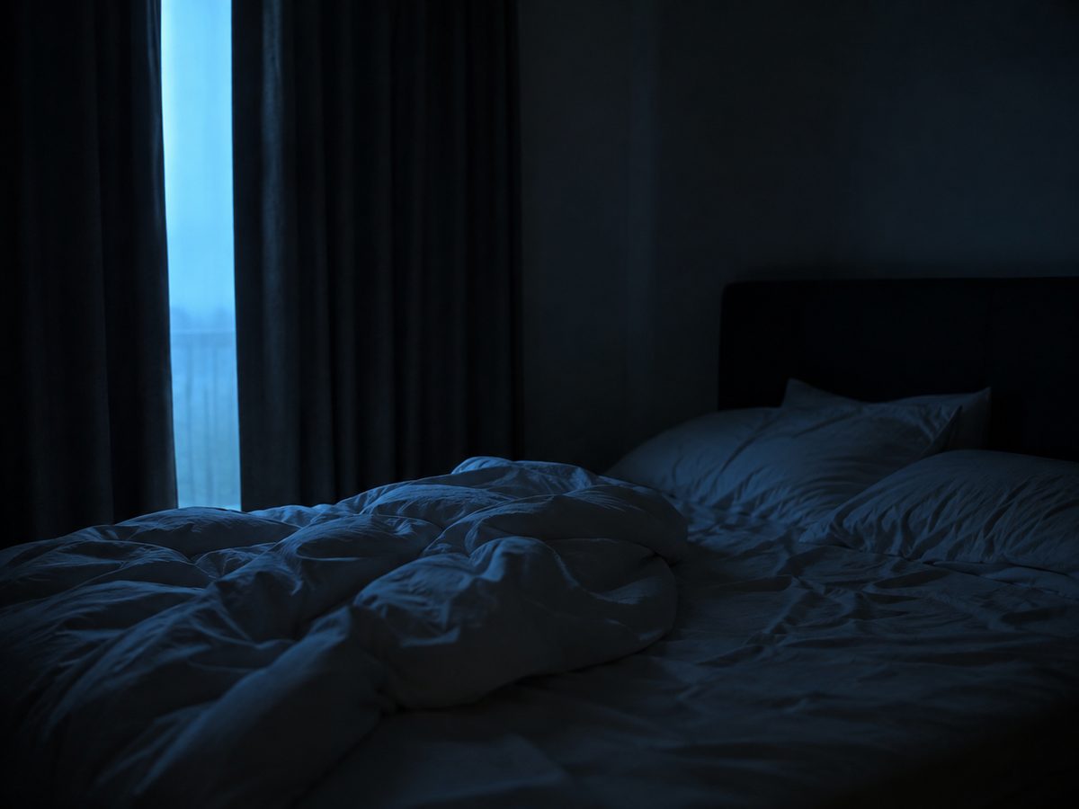 An unmade bed with rumpled white bedding in a dark bedroom, with faint blue pre-dawn light showing through a gap in the curtains