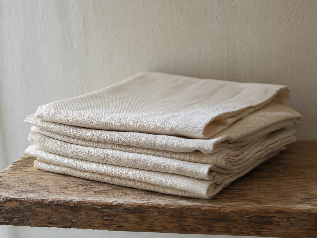 A neatly folded stack of plain undyed natural cotton cloth resting on a weathered wooden shelf in soft daylight